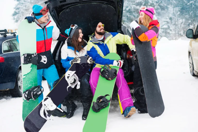 Image is of a group of friends holding snowboards near a car in a snowy resort, enjoying Aspen spring break with luxury winter travel experience